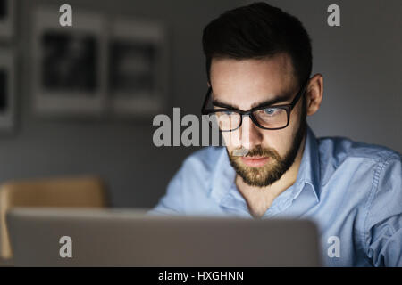 Ritratto di bello uomo barbuto con gli occhiali a lavorare con il computer portatile in camera oscura a tarda notte, il suo viso illuminato dallo schermo Foto Stock