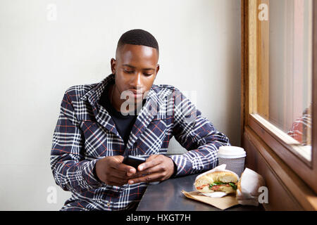 Ritratto di giovane africano seduto a un coffee shop e invio di un messaggio di testo dal proprio telefono cellulare Foto Stock