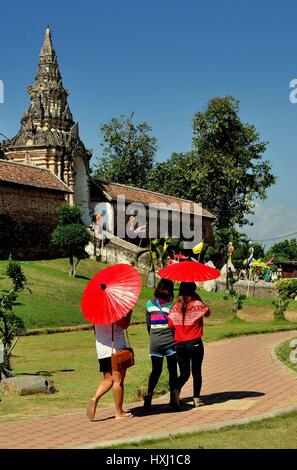 Lampang, Tailandia - 28 dicembre 2012: Tre Tailandese donne ombreggiatura stessi con carta rossa ombrelloni avvicinando l'ingresso murato Wat Phra That L Foto Stock