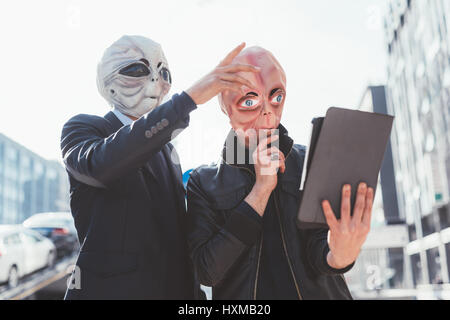 Due uomini che indossano maschere di alieni utilizzando tablet tenere in mano all'aperto nella città di luce posteriore - la strana, tecnologia, concetto di halloween Foto Stock