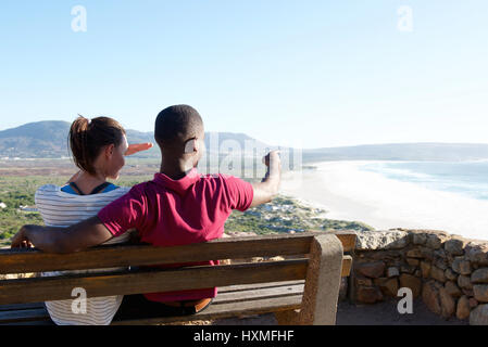 Vista posteriore del Ritratto di giovane coppia seduta su una panchina con uomo puntando verso il mare, una giovane coppia in vacanza. Foto Stock