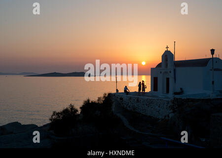 Le persone accanto ad una piccola chiesa sul lungomare potrete ammirare il tramonto a Mykonos, Grecia Foto Stock