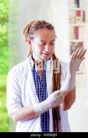 Giovane medico con lunghi dread si blocca in posa per la telecamera mentre mettere dei guanti di gomma, clinica in background, concetto medico Foto Stock