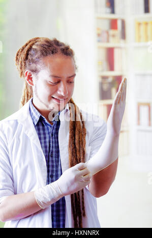 Giovane medico con lunghi dread si blocca in posa per la telecamera mentre mettere dei guanti di gomma, clinica in background, concetto medico Foto Stock