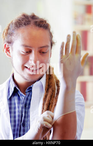 Giovane medico con lunghi dread si blocca in posa per la telecamera mentre mettere dei guanti di gomma, clinica in background, concetto medico Foto Stock