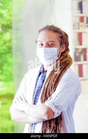 Giovane medico con lunghi dread si blocca in posa per la telecamera, indossa una maschera facciale che copre la bocca, clinica di sfondo, concetto medico Foto Stock