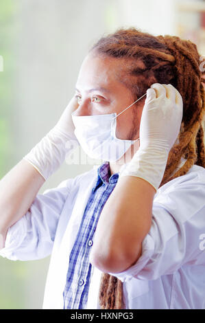 Giovane medico con lunghi dread si blocca in posa per fotocamera, regolazione della maschera facciale che copre la bocca, clinica di sfondo, concetto medico Foto Stock
