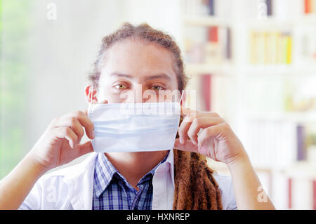 Giovane medico con lunghi dread si blocca in posa per fotocamera, regolazione della maschera facciale che copre la bocca, clinica di sfondo, concetto medico Foto Stock