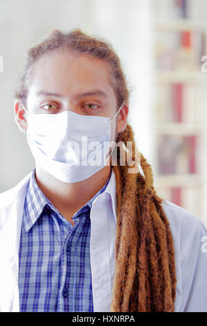 Giovane medico con lunghi dread si blocca in posa per la telecamera, indossa una maschera facciale che copre la bocca, clinica di sfondo, concetto medico Foto Stock