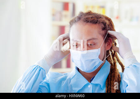 Giovane medico con lunghi dread si blocca in posa per fotocamera, regolazione della maschera facciale che copre la bocca, clinica di sfondo, concetto medico Foto Stock