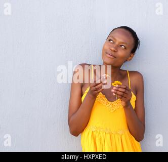 Ritratto di giovane donna africana in abito giallo tenendo un fiore pensando contro il muro grigio Foto Stock