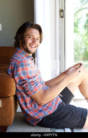 Ritratto di giovane sorridente uomo seduto sul pavimento con il cellulare Foto Stock