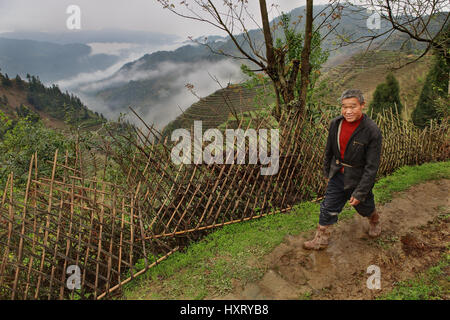 Villaggio di Pingan, provincia di Guangxi, Cina - 5 Aprile 2010: uomo asiatico, indossare stivali di gomma, camminando sulla strada di fango, passato il recinto di vimini sullo sfondo Foto Stock