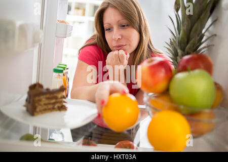 Femmina attraente sulla dieta nel dilemma se mangiare pezzo di torta al cioccolato o arancione dal frigo Foto Stock