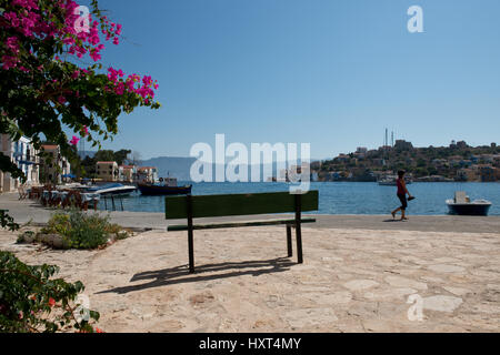 Hafenquai mit Spaziergänger, grüner Banca, Booten, rosa Bougainvillea und bunten Häusern, Insel Kastellorizo, Dodekanes, Griechenland Foto Stock