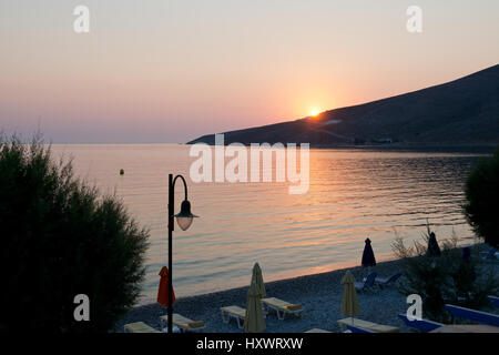 Tramonto sulla baia di Livadia con spiaggia in ciottoli, ombrelloni e lampione in primo piano, isola di Tilos, Dodecaneso, Grecia Foto Stock