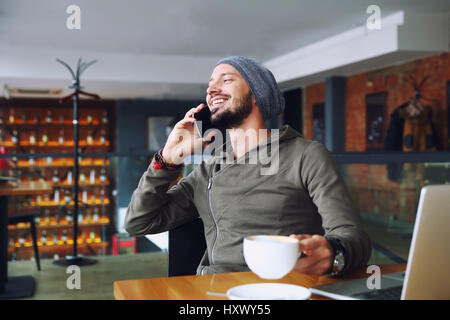 Giovane bello hipster l uomo con la barba in seduta cafe parlando telefono mobile, tenendo tazza di caffè e sorridente. Sul computer portatile sul tavolo di legno. Foto Stock