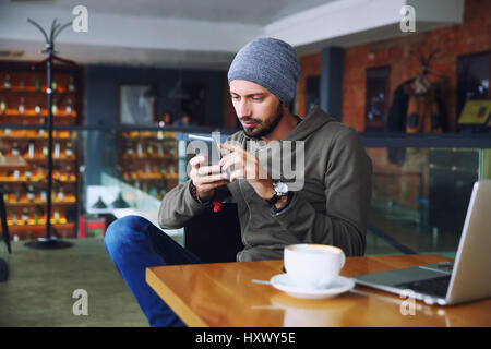 Giovane bello hipster l uomo con la barba in seduta cafe parlando telefono mobile, tenendo tazza di caffè e sorridente. Sul computer portatile sul tavolo di legno. Foto Stock