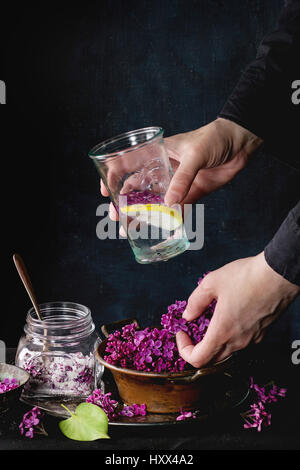 Mani femminili tenere il vetro di limonata lilla con il limone. Il vasetto di vetro di confetti fiori lilla e vintage vaso di fiori freschi sulla tovaglia nero su bl Foto Stock