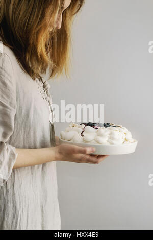 Donna torta di degustazione. In casa torta di meringa Pavlova con panna, zucchero in polvere di mirtilli freschi e salsa di mirtilli in mani femminili su grigio Foto Stock