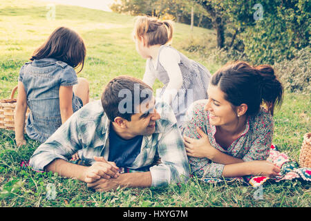 La famiglia felice di quattro giacente in erba in autunno. Il giovane guardando a vicenda con amore come le ragazze giocare. Effetto caldo aggiunto. Foto Stock