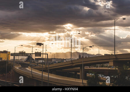 La luce del sole raggiante attraverso le nuvole scure nel deserto di Las Vegas, Nevada. Autostrada cavalcavia, città edifici e montagne distanti su una bella stor Foto Stock