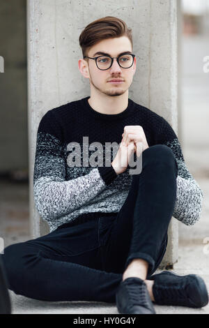 Moda uomo seduto vicino al muro con gli occhiali sulle mani Foto Stock