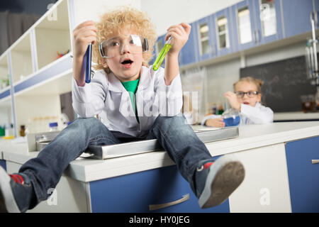 Piccolo Ragazzo seduto nel lavandino nel laboratorio di scienze e la tenuta dei tubi di test con reagenti Foto Stock