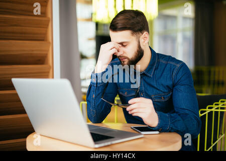 Sensazione di esaurimento. Frustrati giovane uomo barba massaggiando il suo naso e tenere gli occhi chiusi mentre è seduto al suo posto di lavoro in ufficio Foto Stock