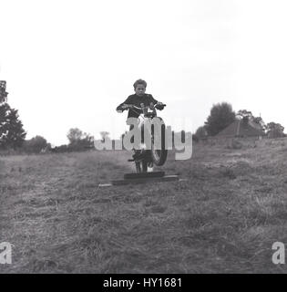 Anni sessanta, storico fanciullo in un campo erboso su una moto motocross praticando la sua tecnica per andare oltre una gobba o ostacolo, Inghilterra. Foto Stock