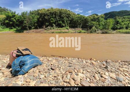 Confezioni di donne di Akha Ya-Er tribù della collina sul sassoso banca del Nam Phak fiume in attesa di attraversare da sponda sinistra-Nam Pak Noy town a destra Foto Stock