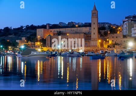 La vista notturna di Marsaskala chiesa parrocchiale di Sant'Anna (St. Anne) oltre il Marsaskala Creek con le tradizionali barche da pesca maltesi (Luzzu). Malta Foto Stock