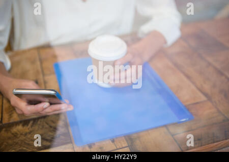 Sezione mediana della donna azienda tazza di caffè e telefono cellulare visto attraverso la finestra cafe Foto Stock