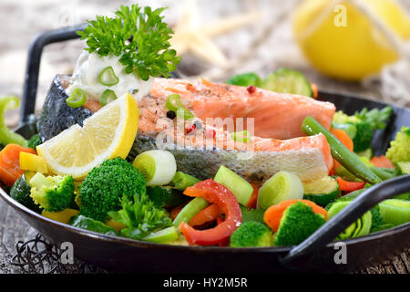 Gustosi piatti fritti e grigliate bistecca di salmone su misto di verdure colorate servita in una padella, limoni e una rete da pesca in background Foto Stock