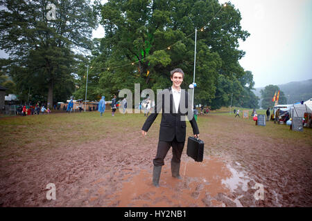 Uomo che indossa un abito nero tenendo un breve caso in piedi in una pozza di fango al Green Man festival, Galles Foto Stock