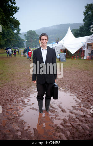 Uomo che indossa un abito nero tenendo un breve caso in piedi in una pozza di fango al Green Man festival, Galles Foto Stock