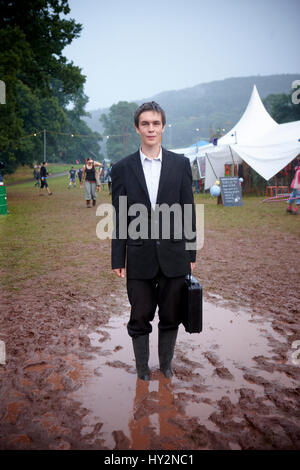 Uomo che indossa un abito nero tenendo un breve caso in piedi in una pozza di fango al Green Man festival, Galles Foto Stock