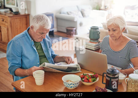 Gli anziani con un computer portatile e la lettura dei giornali a colazione Foto Stock