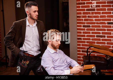 Grande momento in barberia. Giovane uomo barbuto getting haircut dal parrucchiere in giacca marrone Foto Stock