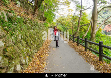 Donna turistico con sciarpa rossa si alza e si affaccia la fotocamera in strada con caduto foglie di autunno nella foresta a Kyoto, Giappone Foto Stock
