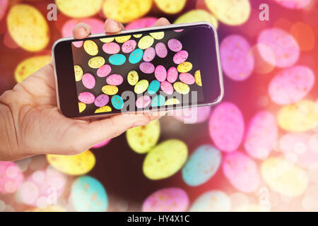Mano che tiene il telefono cellulare contro uno sfondo bianco contro colorate uova di pasqua nel cesto di vimini con fiore Foto Stock