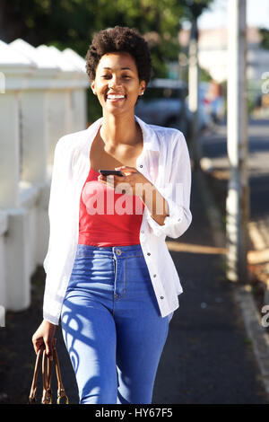 Ritratto di un sorridente giovane donna africana a piedi lungo la strada con il telefono cellulare Foto Stock