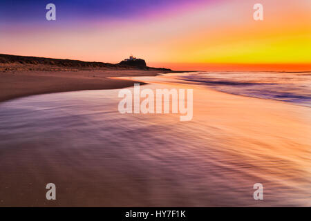 Ampia spiaggia di sabbia sulla testa Nobbys in Newcastle a sunrise. Luminose Rising Sun vernici cielo e oceano onde in arancione sfumature di rosa. Foto Stock