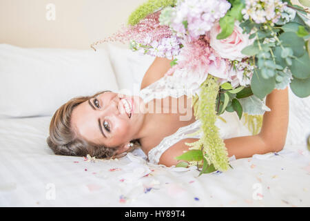 Una sposa la posa sul letto nella sua suite nuziale mentre tiene un mazzo di fiori Foto Stock