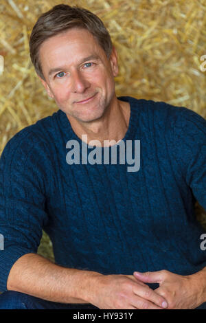 Ritratto di un attraente, di successo e Felice uomo di mezza età maschio indossa un maglione blu seduti su balle di fieno in un granaio o stalle Foto Stock