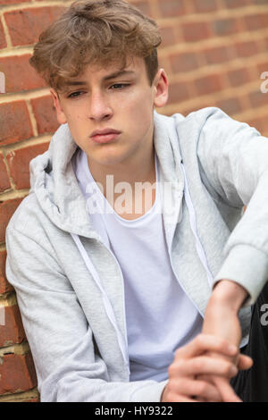 Triste maschio depresso ragazzo adolescente fuori appoggiato su di un muro di mattoni di indossare una felpa con cappuccio grigio Foto Stock