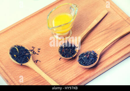 Tè nero con erbe in cucchiai di legno con un contenitore di vetro con il miele su una tavola di legno. Foto Stock