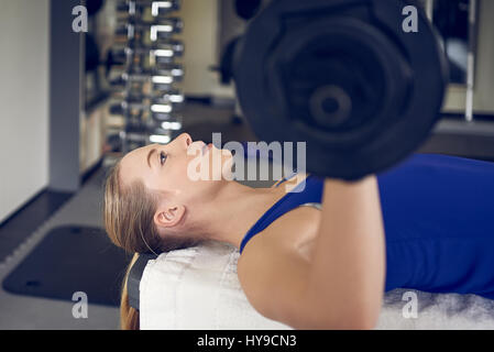 Close-up vista laterale della giovane donna bionda in blu in alto facendo panca esercizio di pressatura con bar-campana in palestra Foto Stock