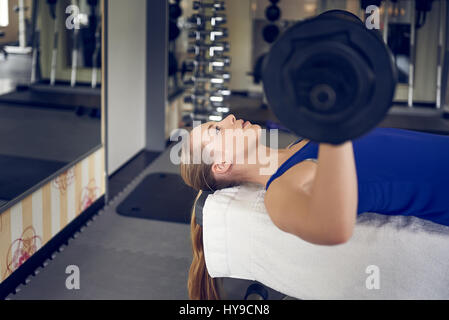 Close-up vista laterale della giovane donna bionda in blu in alto facendo panca esercizio di pressatura con bar-campana in palestra Foto Stock