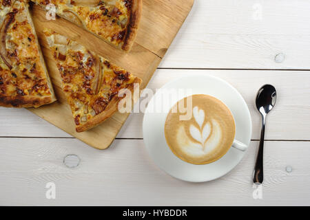 Pezzo di torta su una piastra con una tazza di cappuccino e gli apparecchi in un bianco tavolo di legno. Vista superiore Foto Stock